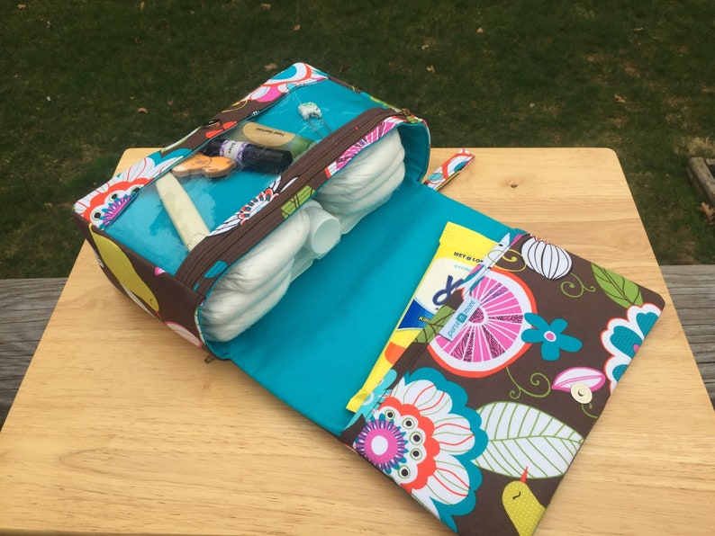 K&ouml;nnte beinhalten: Ge&ouml;ffnete Wickeltasche mit transparentem Deckel, die Windeln und Babyutensilien zeigt. Die Tasche hat ein braunes Blumenmuster mit t&uuml;rkisfarbenen und wei&szlig;en Akzenten. Die Innenseite ist t&uuml;rkis.