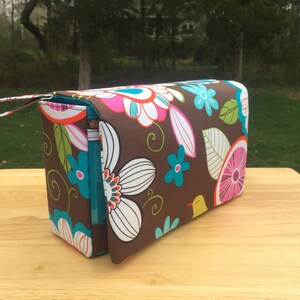 K&ouml;nnte beinhalten: Eine rechteckige braune Kosmetiktasche mit einem bunten Blumenmuster in Pink, Wei&szlig;, T&uuml;rkis und Orange. Die Tasche hat einen t&uuml;rkisfarbenen Rei&szlig;verschluss und einen kleinen Griff. Die Tasche steht auf einer Holzoberfl&auml;che.