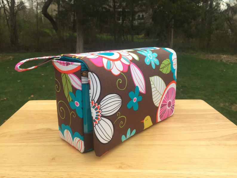 K&ouml;nnte beinhalten: Eine rechteckige braune Kosmetiktasche mit einem bunten Blumenmuster in Pink, Wei&szlig;, T&uuml;rkis und Orange. Die Tasche hat einen t&uuml;rkisfarbenen Rei&szlig;verschluss und einen kleinen Griff. Die Tasche steht auf einer Holzoberfl&auml;che.