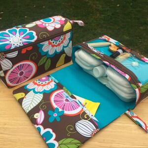 K&ouml;nnte beinhalten: Zwei Wickeltaschen mit einem braunen Blumenmuster mit rosa, t&uuml;rkisfarbenen und wei&szlig;en Blumen. Eine Tasche ist ge&ouml;ffnet und zeigt Windeln und Babyzubeh&ouml;r. Die Taschen haben t&uuml;rkisfarbene Innenr&auml;ume und Rei&szlig;verschl&uuml;sse. Die offene Tasche hat einen Druckknopfverschluss.