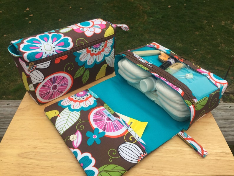 K&ouml;nnte beinhalten: Zwei Wickeltaschen mit einem braunen Blumenmuster mit rosa, t&uuml;rkisfarbenen und wei&szlig;en Blumen. Eine Tasche ist ge&ouml;ffnet und zeigt Windeln und Babyzubeh&ouml;r. Die Taschen haben t&uuml;rkisfarbene Innenr&auml;ume und Rei&szlig;verschl&uuml;sse. Die offene Tasche hat einen Druckknopfverschluss.