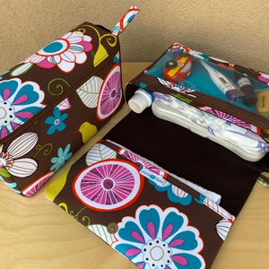 K&ouml;nnte beinhalten: Zwei braune Stoff-Reisetaschen mit einem Blumenmuster in Wei&szlig;, T&uuml;rkis und Rosa. Eine Tasche ist rechteckig mit Rei&szlig;verschluss und Griff. Die andere ist offen und zeigt ein transparentes Fach mit Babyartikeln. Die Taschen liegen auf einer Holzoberfl&auml;che.