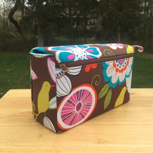 K&ouml;nnte beinhalten: Eine rechteckige braune Stofftasche mit einem farbenfrohen Blumen- und Fruchtmuster. Das Design umfasst gro&szlig;e Bl&uuml;ten in T&uuml;rkis, Wei&szlig; und Rosa, sowie gelbe V&ouml;gel und rosa Zitronenscheiben. Die Tasche hat einen Rei&szlig;verschluss und einen kleinen Griff.
