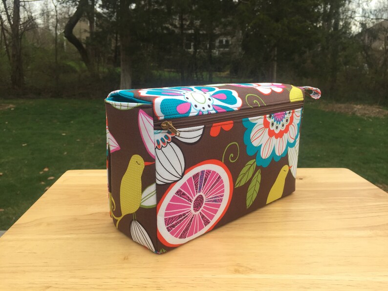 K&ouml;nnte beinhalten: Eine rechteckige braune Stofftasche mit einem farbenfrohen Blumen- und Fruchtmuster. Das Design umfasst gro&szlig;e Bl&uuml;ten in T&uuml;rkis, Wei&szlig; und Rosa, sowie gelbe V&ouml;gel und rosa Zitronenscheiben. Die Tasche hat einen Rei&szlig;verschluss und einen kleinen Griff.