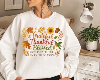 以下が含まれることがあります： 秋をテーマにしたデザインの白いスウェットシャツ。「Grateful, Thankful, Blessed」の文字がオレンジ、赤、緑色で、葉、ヒマワリ、ドングリ、緑色の鳥で囲まれています。「GOD IS FAITHFUL IN EVERY SEASON」の文字も含まれています。