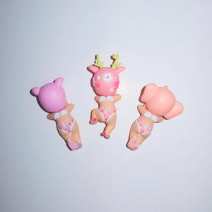 K&ouml;nnte beinhalten: Drei skurrile Figuren, jede mit einem anderen Tierkopf: ein B&auml;r, ein Reh und ein Elefant. Sie alle tragen wei&szlig;e Bikinis mit floralem Muster. Die Figuren sind rosa und hellbraun, vor wei&szlig;em Hintergrund.