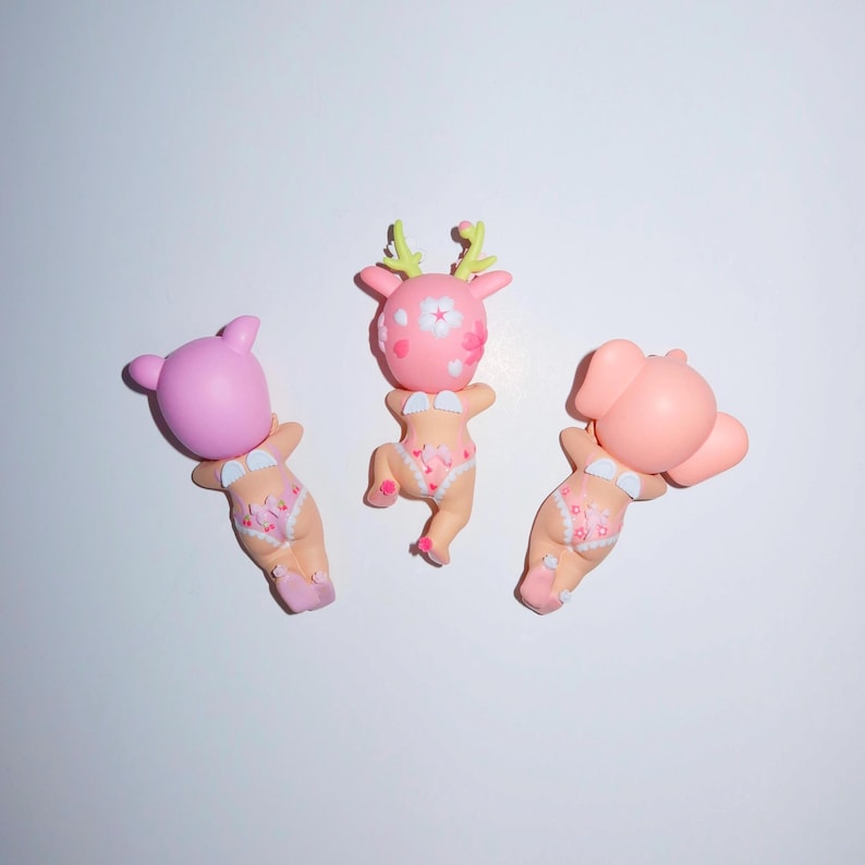 K&ouml;nnte beinhalten: Drei skurrile Figuren, jede mit einem anderen Tierkopf: ein B&auml;r, ein Reh und ein Elefant. Sie alle tragen wei&szlig;e Bikinis mit floralem Muster. Die Figuren sind rosa und hellbraun, vor wei&szlig;em Hintergrund.