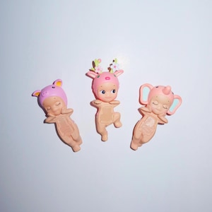 K&ouml;nnte beinhalten: Drei kleine Figuren, jede mit einem Hut mit Tiermotiv: ein Schwein, ein Reh und ein Elefant. Die Figuren sind hellrosa und beige, mit geschlossenen Augen und verschr&auml;nkten Armen.
