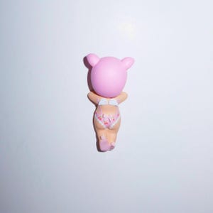 K&ouml;nnte beinhalten: Eine rosa Figur eines Babys mit b&auml;ren&auml;hnlichem Kopf, das einen rosa und wei&szlig; gebl&uuml;mten Windel tr&auml;gt. Die Figur wird von hinten betrachtet, mit erhobenen Armen. Der Hintergrund ist schlicht wei&szlig;.