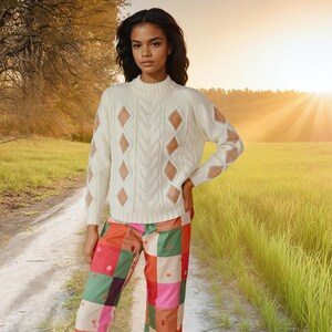 May include: A cream cable-knit jumper with diamond cut-outs on the sleeves and chest. The jumper is worn with patchwork trousers in various colours. The model is standing on a dirt track in a field.