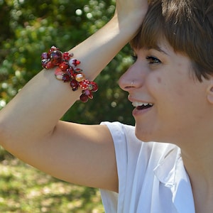 May include: A cluster bracelet with red, white, and clear beads. The bracelet features various shapes and sizes of beads, including round, oval, and irregular forms. The beads are arranged in a cascading design, creating a textured and eye-catching accessory.