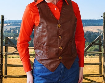May include: A brown leather vest with a red button-down shirt underneath. The vest has a pointed collar and multiple pockets. The person is wearing blue jeans.