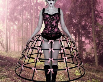 Puede incluir: Un conjunto de inspiración gótica con un corsé rosa y negro, una falda jaula adornada con lazos rosas y botas negras. La modelo tiene la piel pálida y un maquillaje dramático, sobre un fondo de bosque rosa.