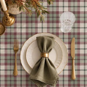 May include: A table setting featuring a plaid tablecloth with maroon, sage green, and white stripes. A white plate with a gold rim holds an olive green napkin secured by a gold ring. A gold fork and knife, a glass, and a candle are present.