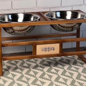 Peut inclure: Un support surélevé en bois pour animaux avec deux gamelles en acier inoxydable. La structure est en bois teinté marron et comporte une plaque centrale rectangulaire avec le nom Meeko gravé en écriture cursive foncée sur un panneau clair.