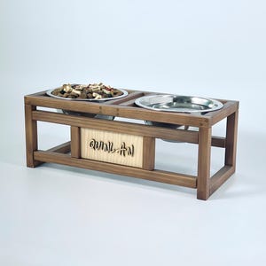 Puede incluir: Un comedero de madera para mascotas con dos cuencos de acero inoxidable. Un cuenco contiene comida para mascotas. El comedero tiene un diseño rectangular con el nombre "QUINLAN" tallado en la parte delantera. La madera tiene un tono marrón cálido.