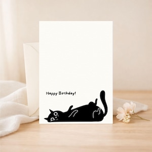 Puede incluir: Una tarjeta de felicitación blanca con una ilustración negra de un gato tumbado boca arriba. Las palabras "Happy Birthday!" están impresas encima del gato. Un sobre blanquecino y un pequeño ramo de flores están junto a la tarjeta.