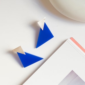 Op de afbeelding: Een paar oorknopjes met een felblauw driehoekig acrylonderdeel bevestigd aan een kleiner zilverkleurig driehoekig metalen stuk. De oorbellen hebben een geometrisch ontwerp met scherpe hoeken en een gladde, gepolijste afwerking op het metaal.