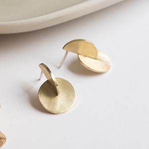 May include: A pair of gold-toned stud earrings featuring a circular base with a perpendicular semi-circular piece attached to the front. The metal has a brushed finish and includes thin silver-toned posts for attachment to the earlobe.