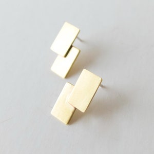 Puede incluir: Dos pares de pendientes de botón rectangulares dorados. Cada pendiente presenta dos rectángulos superpuestos. Los pendientes se muestran sobre una superficie gris claro, mostrando su diseño minimalista y acabado metálico.