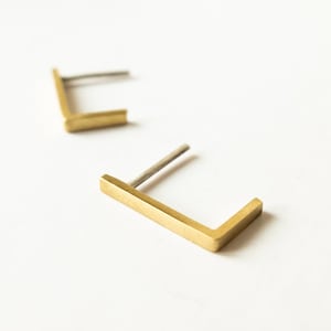 Puede incluir: Dos pendientes minimalistas en forma de L de color dorado. Cada pendiente tiene un poste recto para insertar y un diseño en ángulo recto. Los pendientes están sobre un fondo blanco, resaltando su forma geométrica simple.