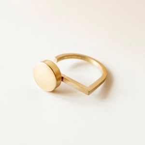 Könnte beinhalten: Ein goldener Ring mit geometrischem Design. Der Ring hat eine runde Scheibe, die an einer rechteckigen Stange befestigt ist.