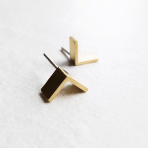 Op de afbeelding: Een paar goudkleurige, L-vormige oorstekers. Elke oorbel heeft een pin voor inbrenging in het oor. De oorbellen worden gepresenteerd op een wit oppervlak, met één oorbel scherp en de andere licht wazig op de achtergrond.