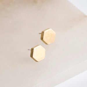 Könnte beinhalten: Ein Paar goldfarbene, sechseckige Ohrstecker. Die Ohrringe haben eine gebürstete Metalloberfläche und sind vor einem weißen Hintergrund platziert. Das Design der Ohrringe ist minimalistisch.