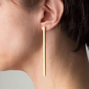 Può includere: Un lungo e sottile orecchino a barra color oro con un profilo rettangolare liscio. L'orecchino pende verticalmente dal lobo dell'orecchio, caratterizzato da un design pulito e dritto che si estende verso il basso in una forma geometrica.