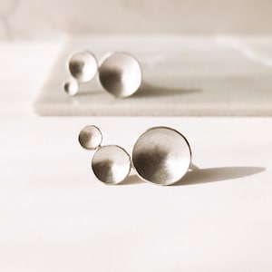 Pode incluir: Um conjunto de duas alianças de prata com um design único que apresenta três círculos sobrepostos. As alianças são feitas de metal liso e polido e têm uma estética moderna e minimalista.
