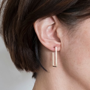 May include: A close-up of a person's ear wearing gold rectangular earrings. The earrings are a simple, modern design, with a straight bar that extends down from the earlobe. The earrings are a warm gold color and appear to be made of metal.