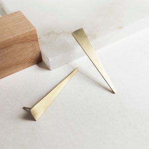 May include: A pair of gold-coloured, triangular earrings. The earrings have a long, slender shape and a minimalist design. They are set against a white background, with a wooden block and a marble slab visible.
