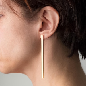 Può includere: Primo piano di un orecchio con un lungo orecchino a barra dorata. L'orecchino ha un design semplice a linea retta, appeso verticalmente. Lo sfondo è di tono neutro, evidenziando lo stile minimalista dell'orecchino.