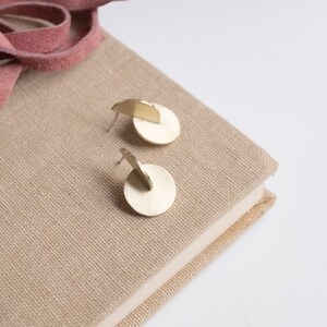 May include: A pair of gold-toned stud earrings, each featuring a flat circular disc base with a smaller semi-circular piece attached perpendicularly to the top, creating a layered geometric structure with a smooth, metallic finish.