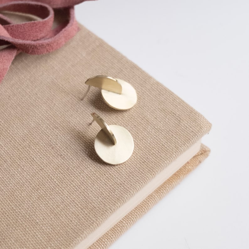 May include: A pair of gold-toned stud earrings, each featuring a flat circular disc base with a smaller semi-circular piece attached perpendicularly to the top, creating a layered geometric structure with a smooth, metallic finish.