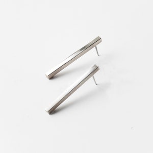 Pode incluir: Um par de brincos de barra prateados. Os brincos são feitos de uma barra retangular de metal com uma borda ligeiramente arredondada. Os brincos são simples e de design moderno.