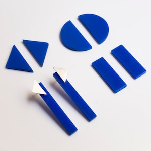 Puede incluir: Varios componentes de joyería geométrica en azul brillante. Las formas incluyen triángulos, rectángulos y semicírculos. Dos pendientes presentan detalles triangulares plateados. Los componentes están dispuestos sobre una superficie blanca.