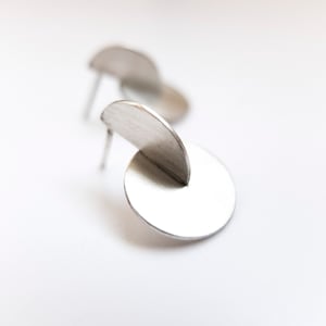 Könnte beinhalten: Ein Paar silberne Ohrringe mit geometrischem Design. Die Ohrringe bestehen aus zwei Kreisen, einer kleiner als der andere, die durch einen dünnen Metallstreifen verbunden sind.