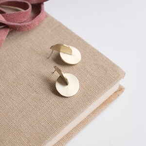 May include: A pair of gold-toned metal stud earrings, each featuring a flat circular base with a perpendicular semi-circular piece attached to the center. The earrings have a brushed finish and are designed with a post-back closure for secure wear.
