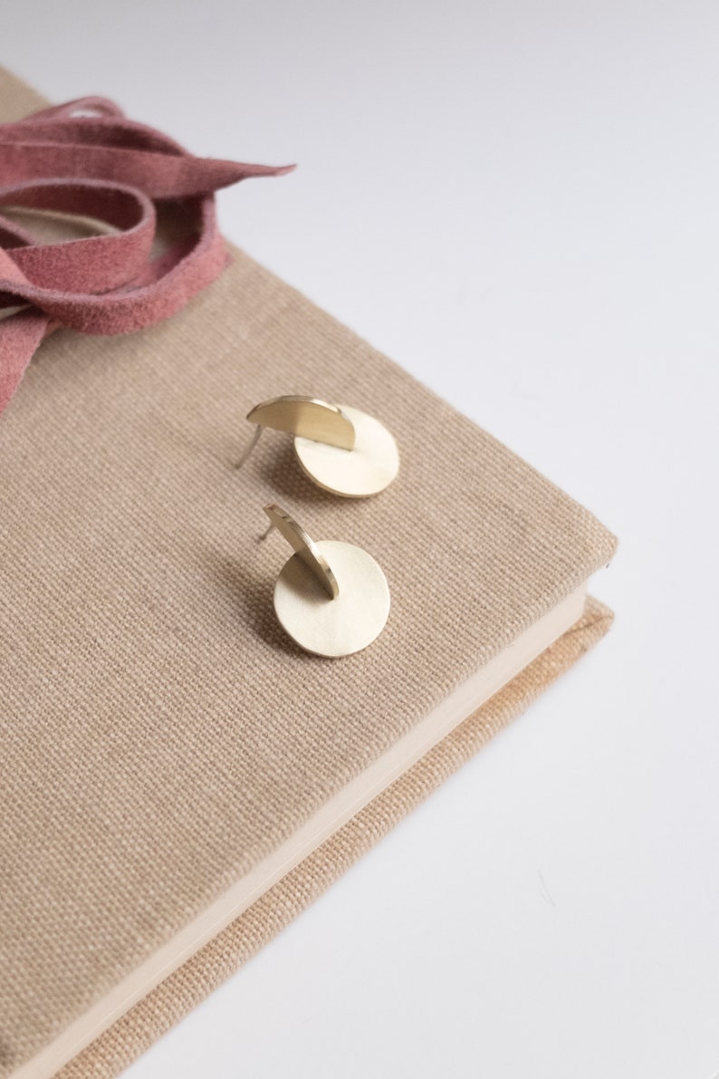 May include: A pair of gold-toned metal stud earrings, each featuring a flat circular base with a perpendicular semi-circular piece attached to the center. The earrings have a brushed finish and are designed with a post-back closure for secure wear.