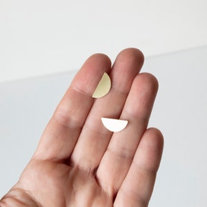 Puede incluir: Dos pendientes pequeños en forma de semicírculo, de color dorado claro. Los pendientes se muestran en la mano de una persona. Diseño sencillo y minimalista.