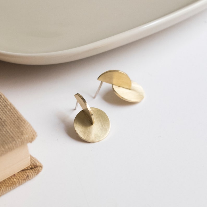 May include: A pair of gold-toned stud earrings featuring a circular base with a perpendicular semi-circular piece attached to the front. The metal has a brushed finish and includes thin silver-toned posts for attachment to the earlobe.