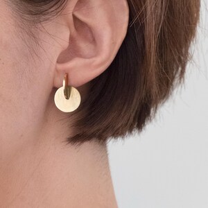 May include: A gold-toned earring featuring a small, polished circular disc suspended from a thin, vertical metal bar. The earring is designed with a smooth, reflective surface and a simple, geometric construction that hangs directly from the earlobe.
