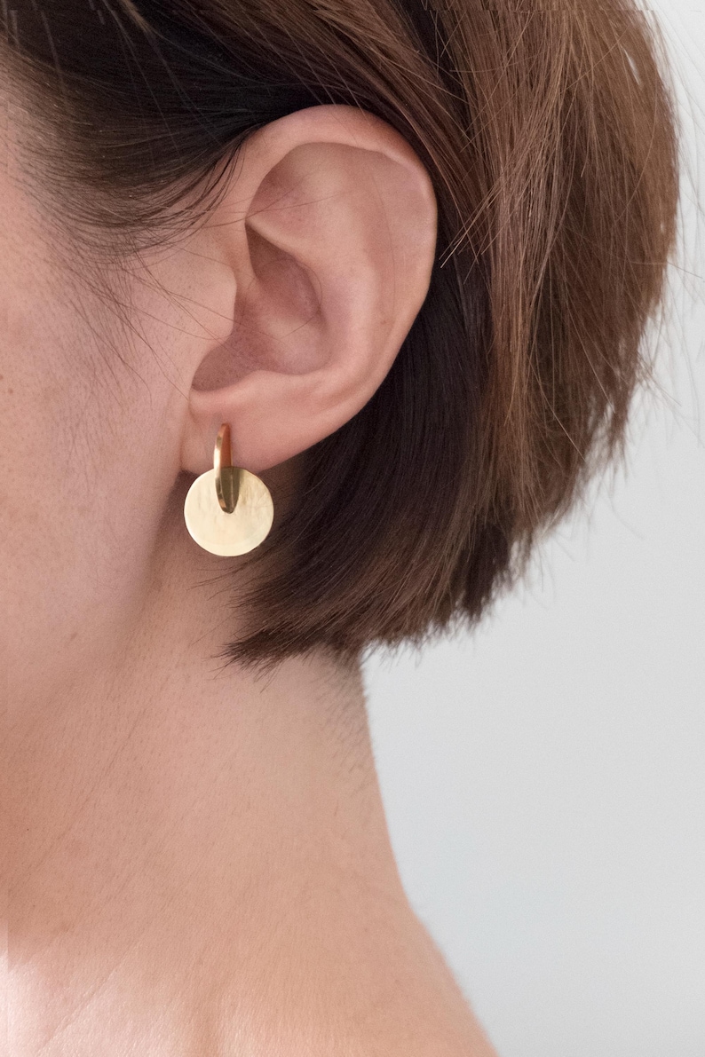 May include: A gold-toned earring featuring a small, polished circular disc suspended from a thin, vertical metal bar. The earring is designed with a smooth, reflective surface and a simple, geometric construction that hangs directly from the earlobe.