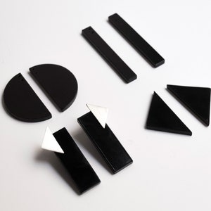 Puede incluir: Un conjunto de pendientes geom&eacute;tricos negros, que incluyen c&iacute;rculos, tri&aacute;ngulos y rect&aacute;ngulos. Algunos pendientes tienen un acento triangular plateado.