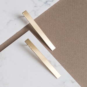 Könnte beinhalten: Zwei goldfarbene Ohrstecker in Form von Stäben werden auf einer weißen Marmoroberfläche vor einem neutralen braunen Hintergrund präsentiert. Die Ohrringe sind rechteckig und haben ein schlichtes, minimalistisches Design. Die Ohrringe sind etwa 5 cm lang.