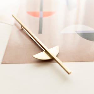 Può includere: Un fermaglio per capelli in metallo dorato con un design geometrico semplice. Il fermaglio presenta una lunga barra dritta con una forma semicircolare all'estremità.