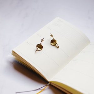 K&ouml;nnte beinhalten: Ein Paar goldfarbene Ohrringe mit minimalistischem Design. Jeder Ohrring hat einen kleinen, flachen Kreis mit einem geraden Stab, der sich davon erstreckt, und einen kleineren Stab in einem Winkel. Die Ohrringe liegen auf einem offenen Notizbuch.