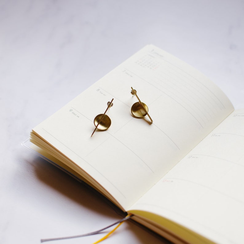 K&ouml;nnte beinhalten: Ein Paar goldfarbene Ohrringe mit minimalistischem Design. Jeder Ohrring hat einen kleinen, flachen Kreis mit einem geraden Stab, der sich davon erstreckt, und einen kleineren Stab in einem Winkel. Die Ohrringe liegen auf einem offenen Notizbuch.