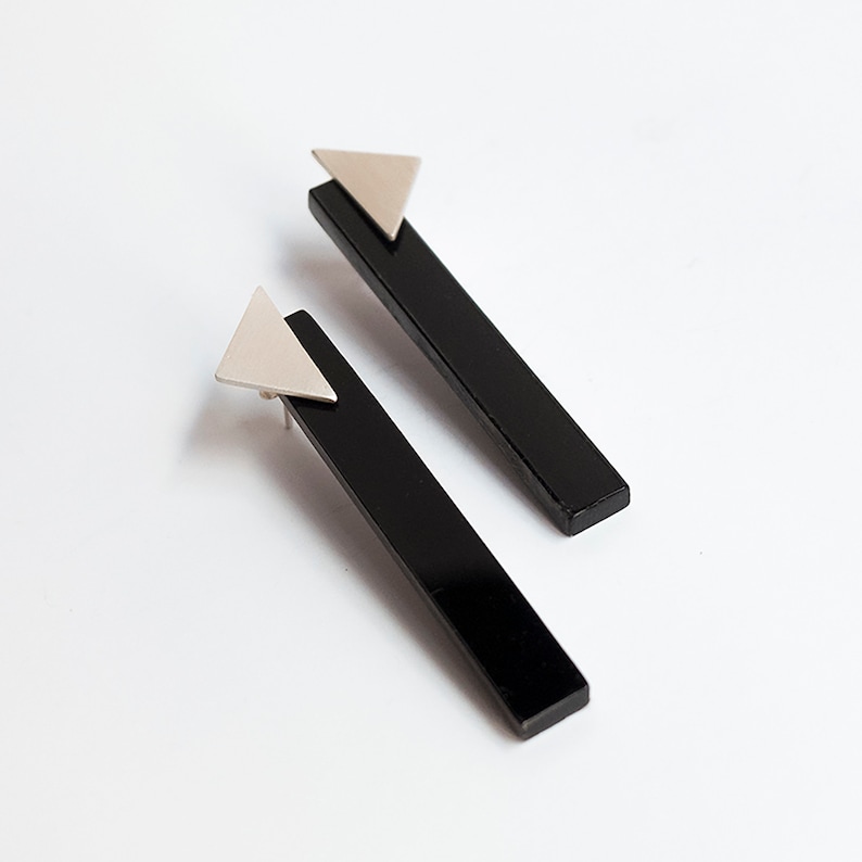 Puede incluir: Un par de pendientes negros y plateados. Cada pendiente tiene una pieza rectangular negra larga con un tri&aacute;ngulo plateado en la parte superior.